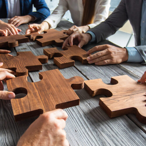 Business people team sitting around meeting table and assembling wooden jigsaw puzzle pieces unity cooperation ideas concept